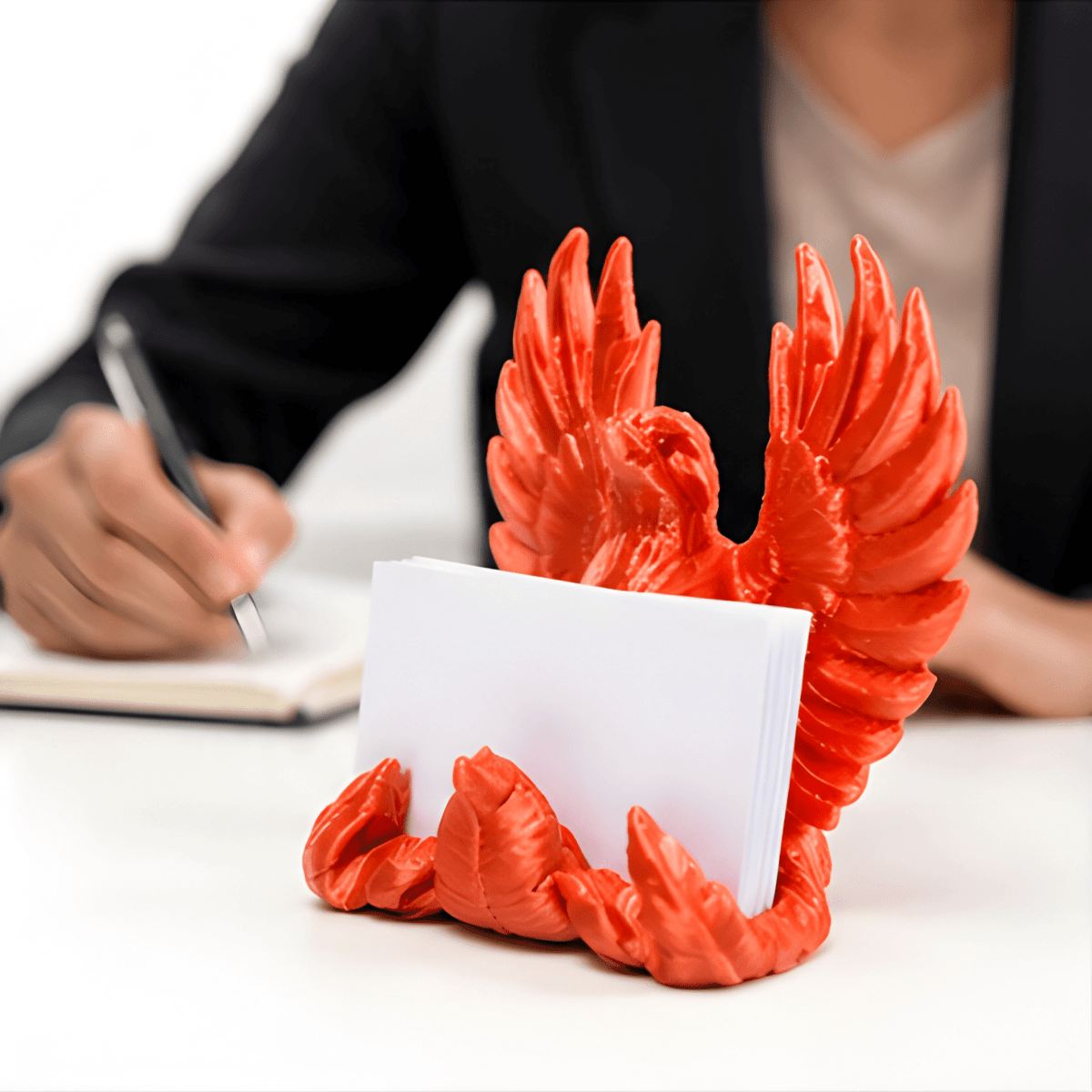 3D-Printed Business Card Holder - SearchPify - Red dragon-shaped card holder with a card on a desk with a person writing.