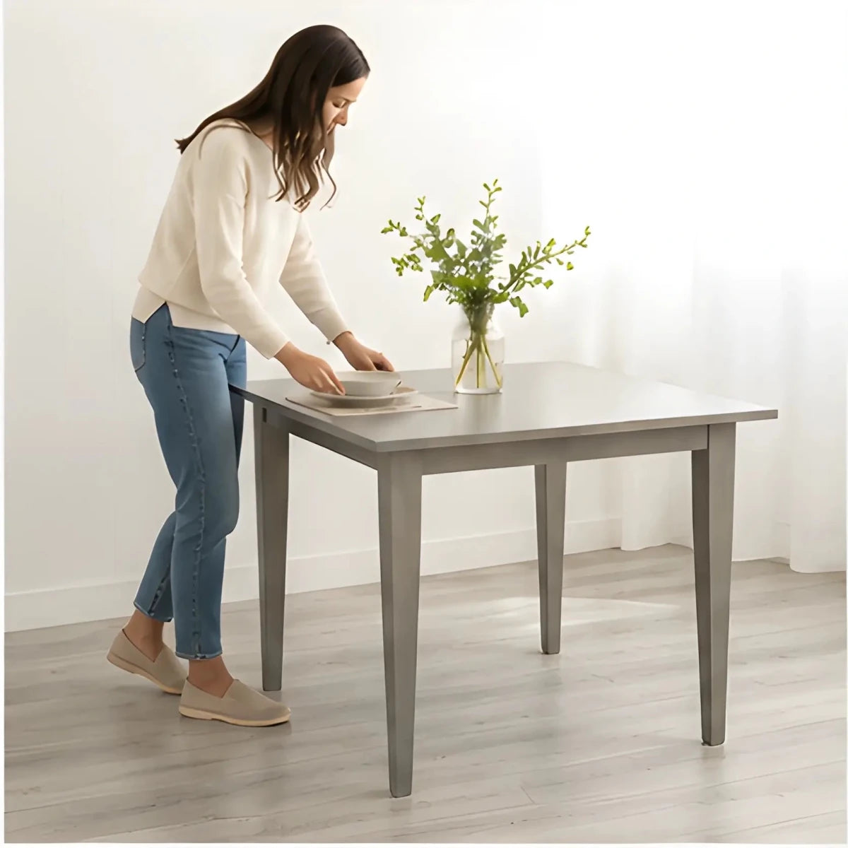The Windermere Square Dining Table - SearchPify - Woman setting a table with a vase of flowers in a minimalistic room.