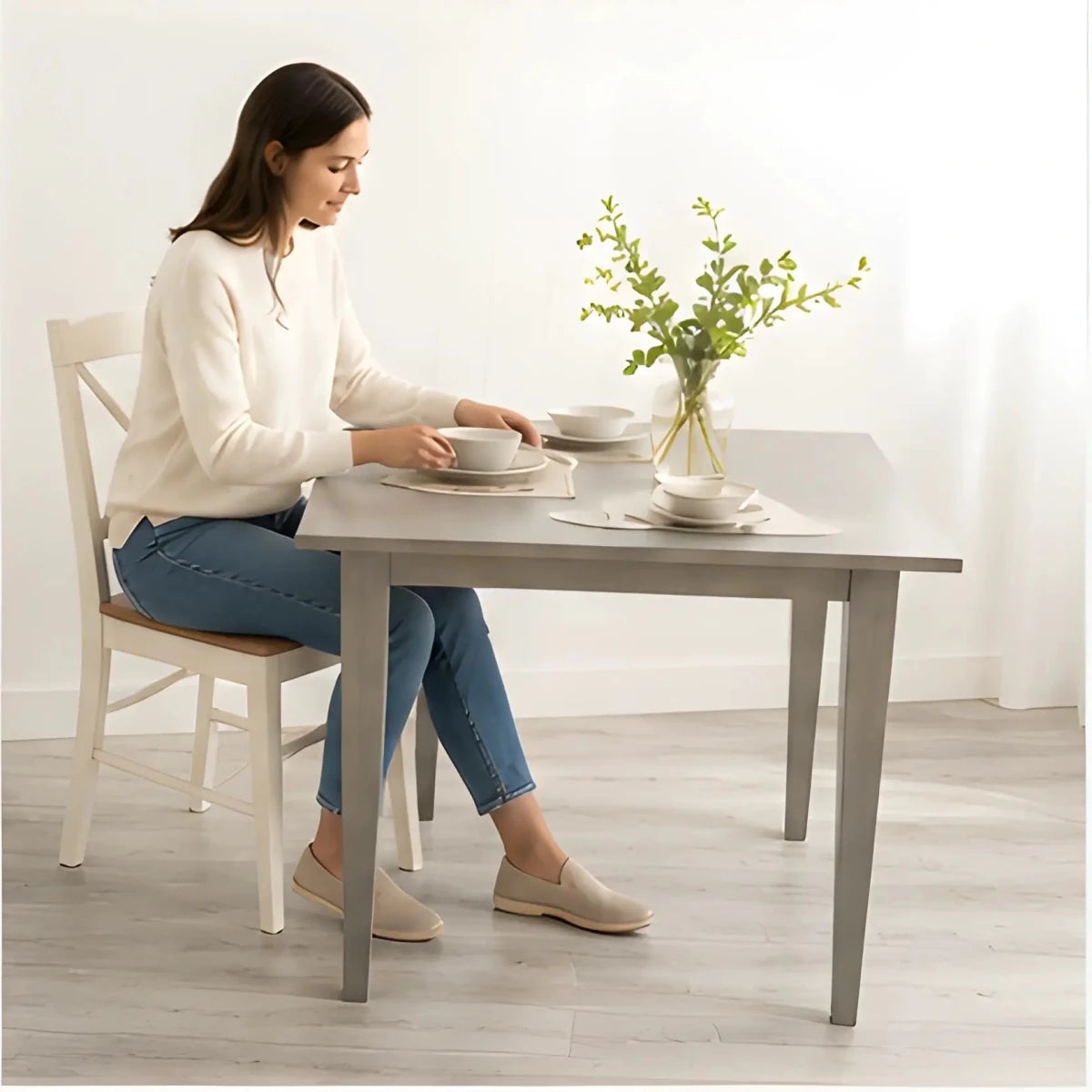 The Windermere Square Dining Table - SearchPify - Woman sitting at a dining table setting plates with a vase of flowers on the table.